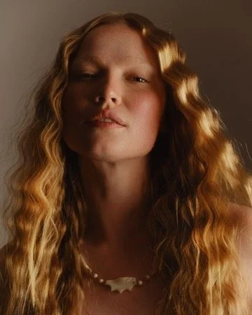 A woman with long, wavy red hair and a confident expression, wearing a necklace made of small white objects. Hair and makeup by Amber Rose.