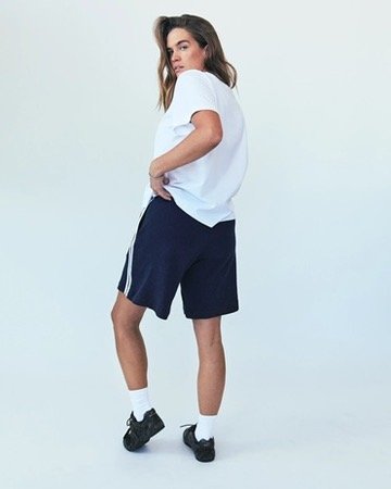 Young woman standing with hands on hips, wearing a white t-shirt, navy shorts, white socks, and black sneakers, against a plain light background. Hair and makeup by Amber Rose.