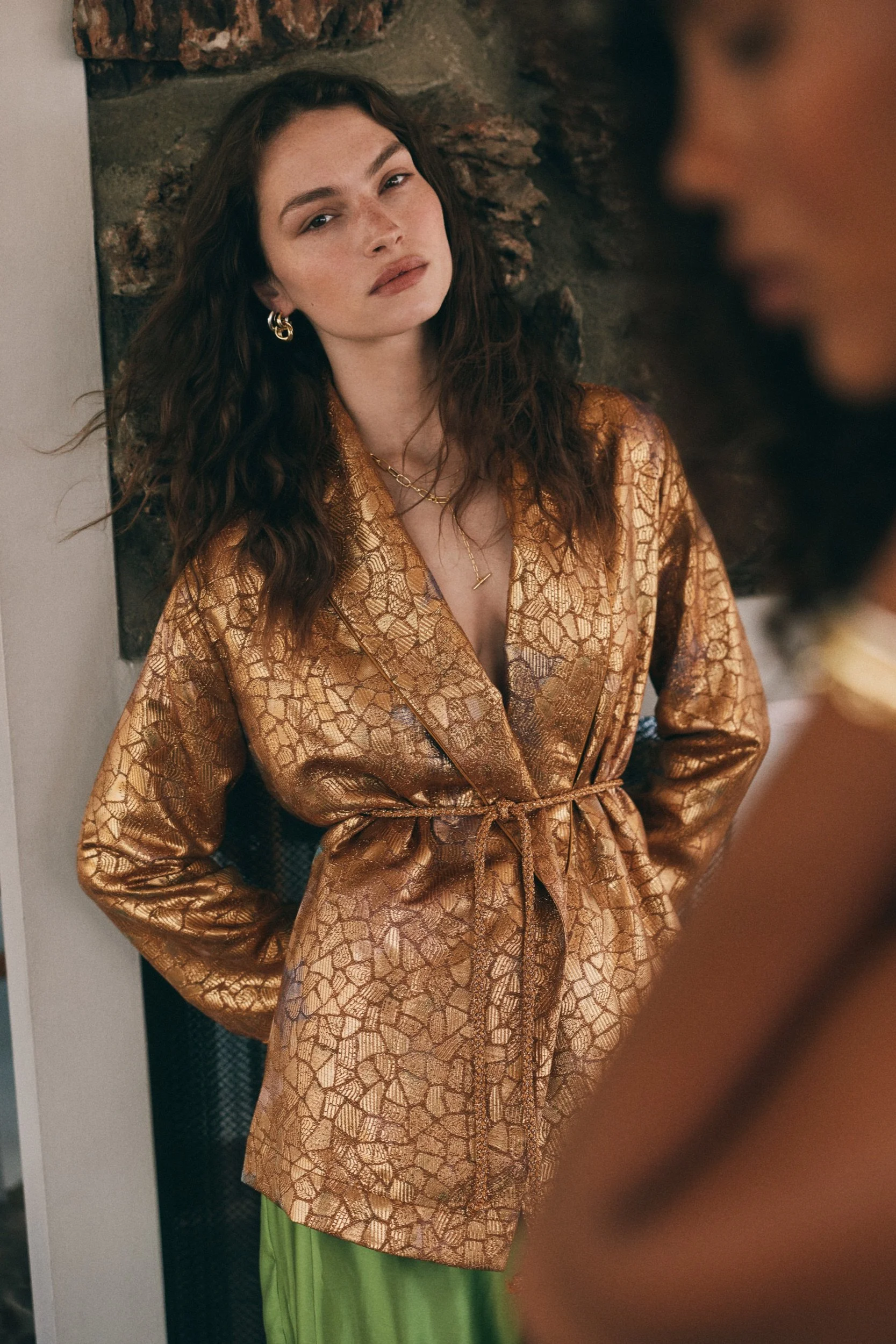 A woman wearing a gold patterned silk robe and a lime green skirt, with jewelry including earrings, necklaces, and rings, standing against a stone wall, with a blurred person on the right side of the image.