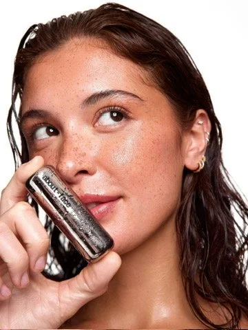 A woman with curly hair and earrings holds a small cosmetics bottle near her face, looking to the side with a thoughtful expression. Hair and makeup by Amber Rose. 