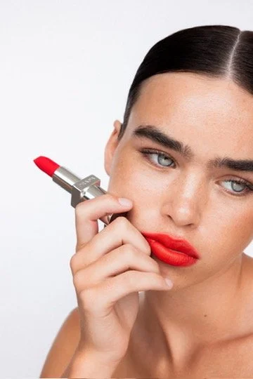 A woman with dark hair applying bright red lipstick with a silver lipstick tube against a plain white background. Hair and makeup by Amber Rose.