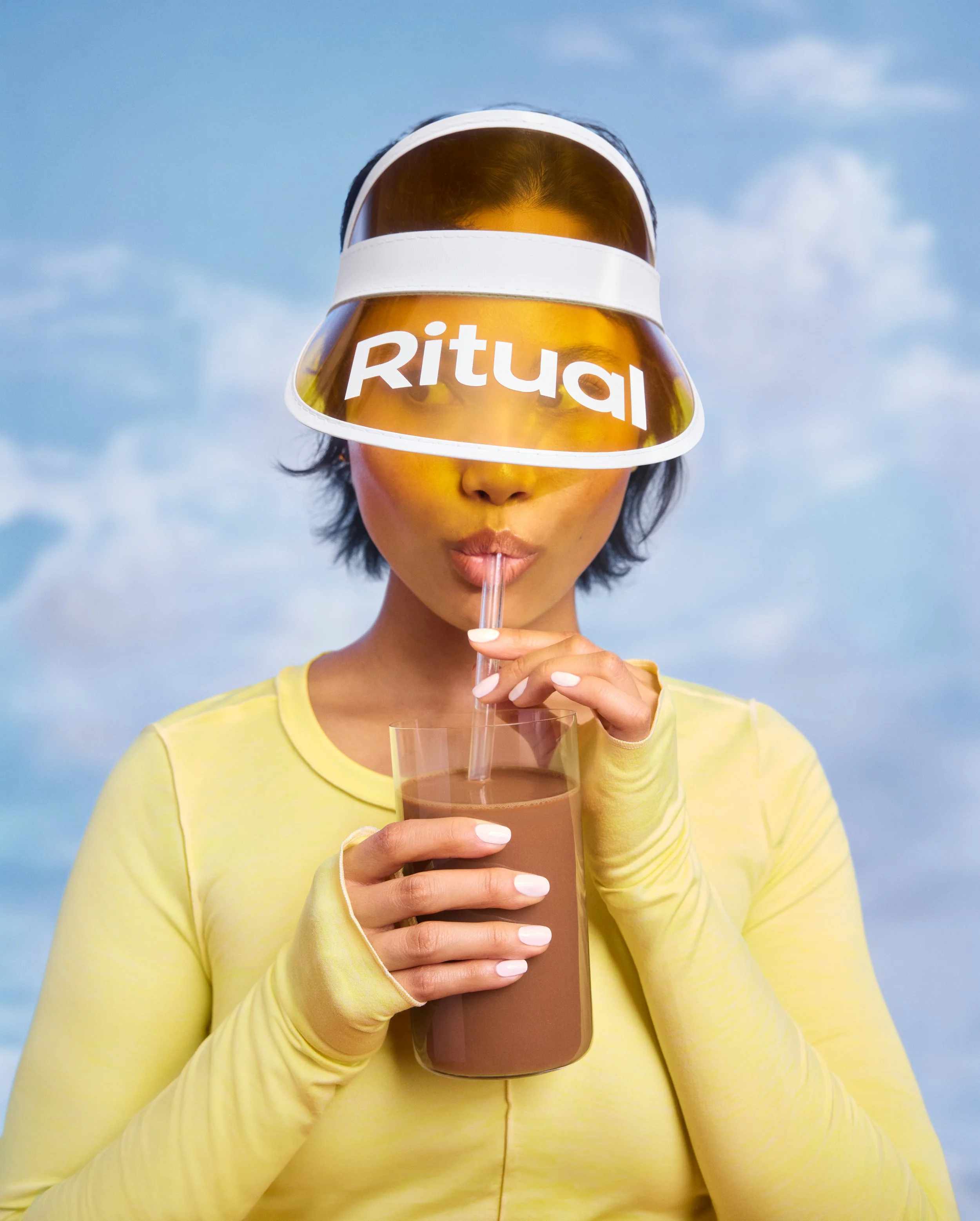 A woman in a yellow top wearing a white visor with a reflective mirror, with the word 'Ritual' written on it, drinking a chocolate beverage through a straw against a blue sky with clouds.