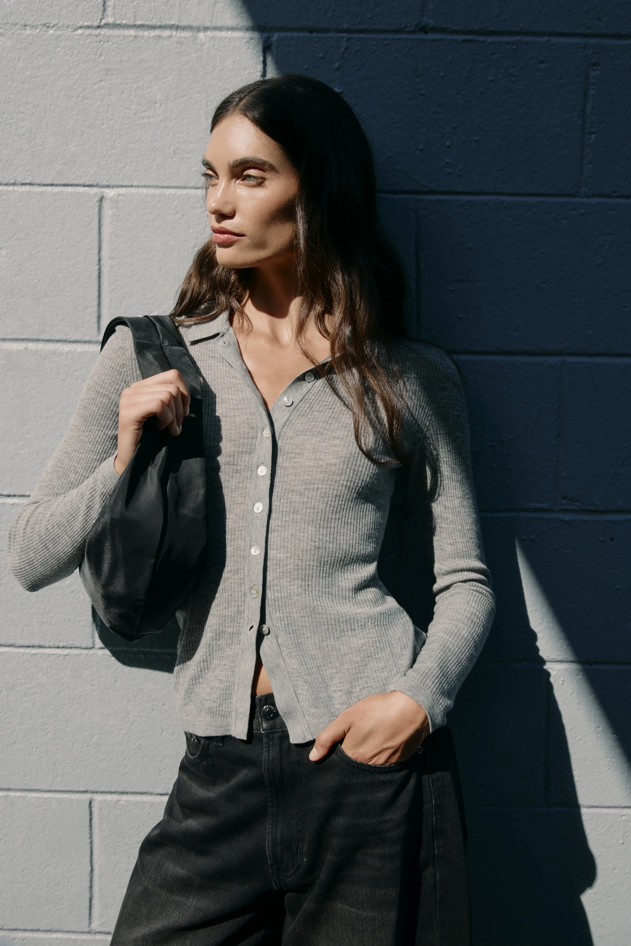 Woman with long brown hair wearing a gray button-up shirt, black jeans, and a black backpack, standing against a two-tone colored wall with shadows cast on her.