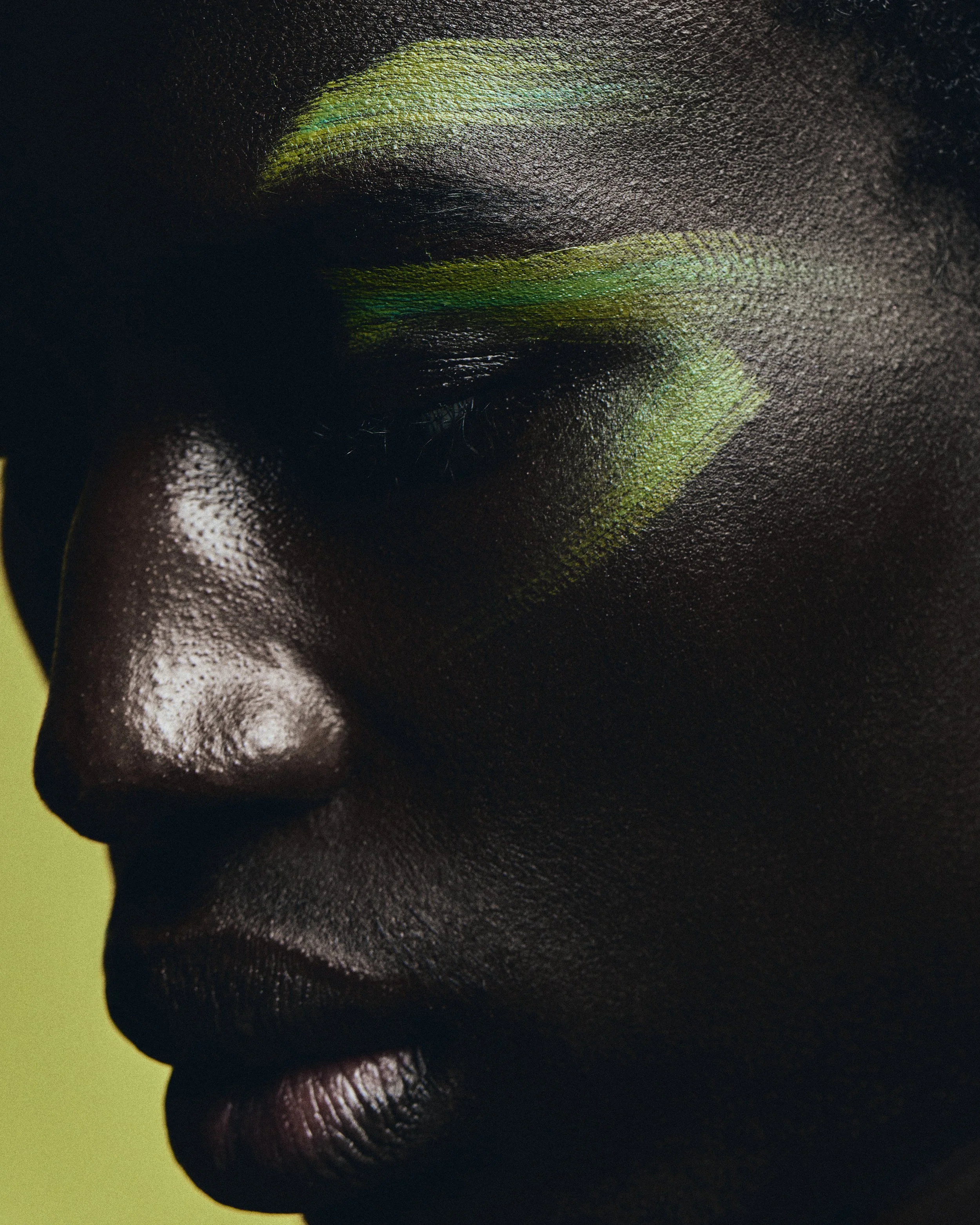 Close-up of a person's face with vibrant green and yellow eye makeup against a dark background.