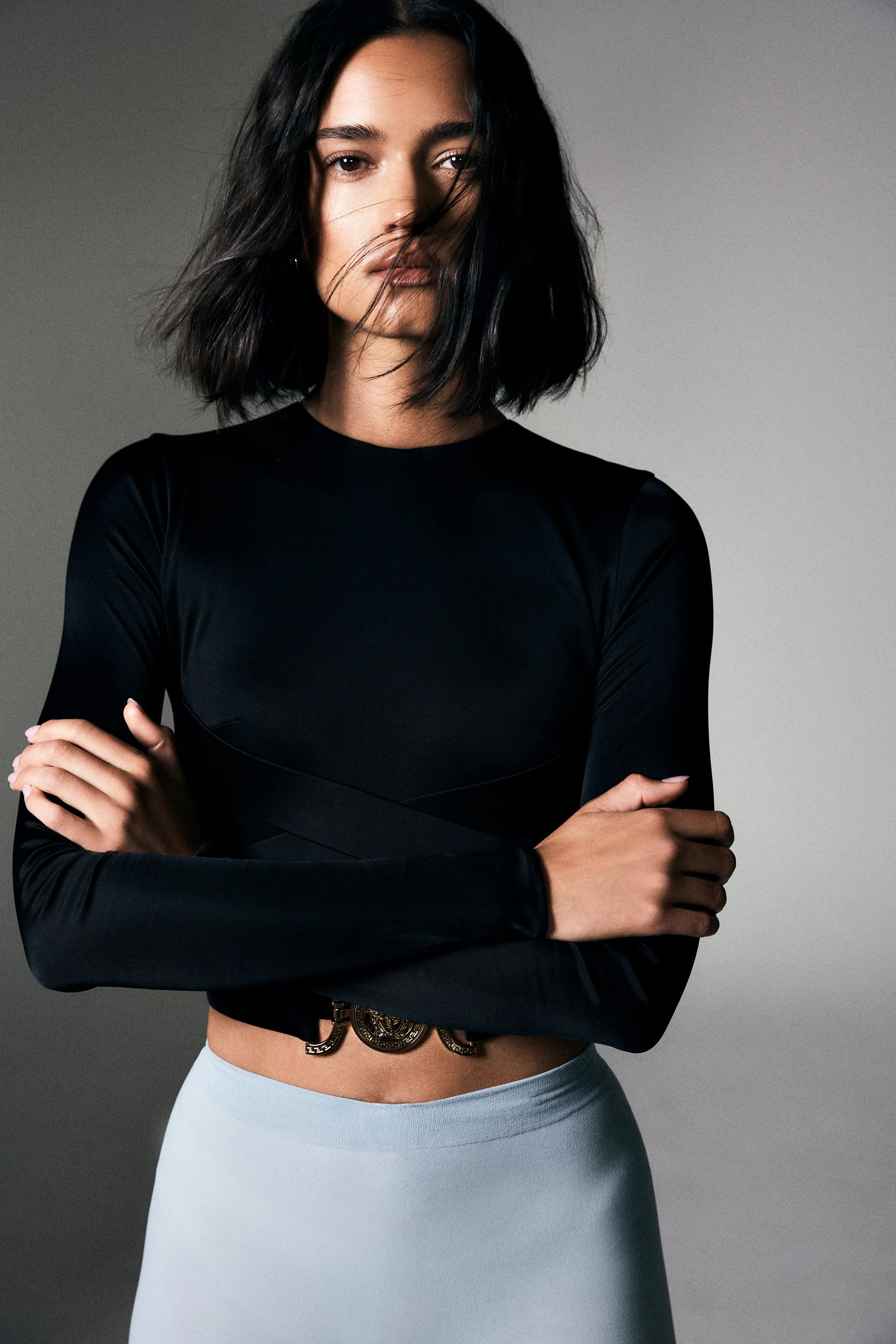 A woman with black hair wearing a black long-sleeve top with crossed arms, a metallic belt, and a light blue skirt, standing against a gray background. Hair and makeup by Amber Rose.