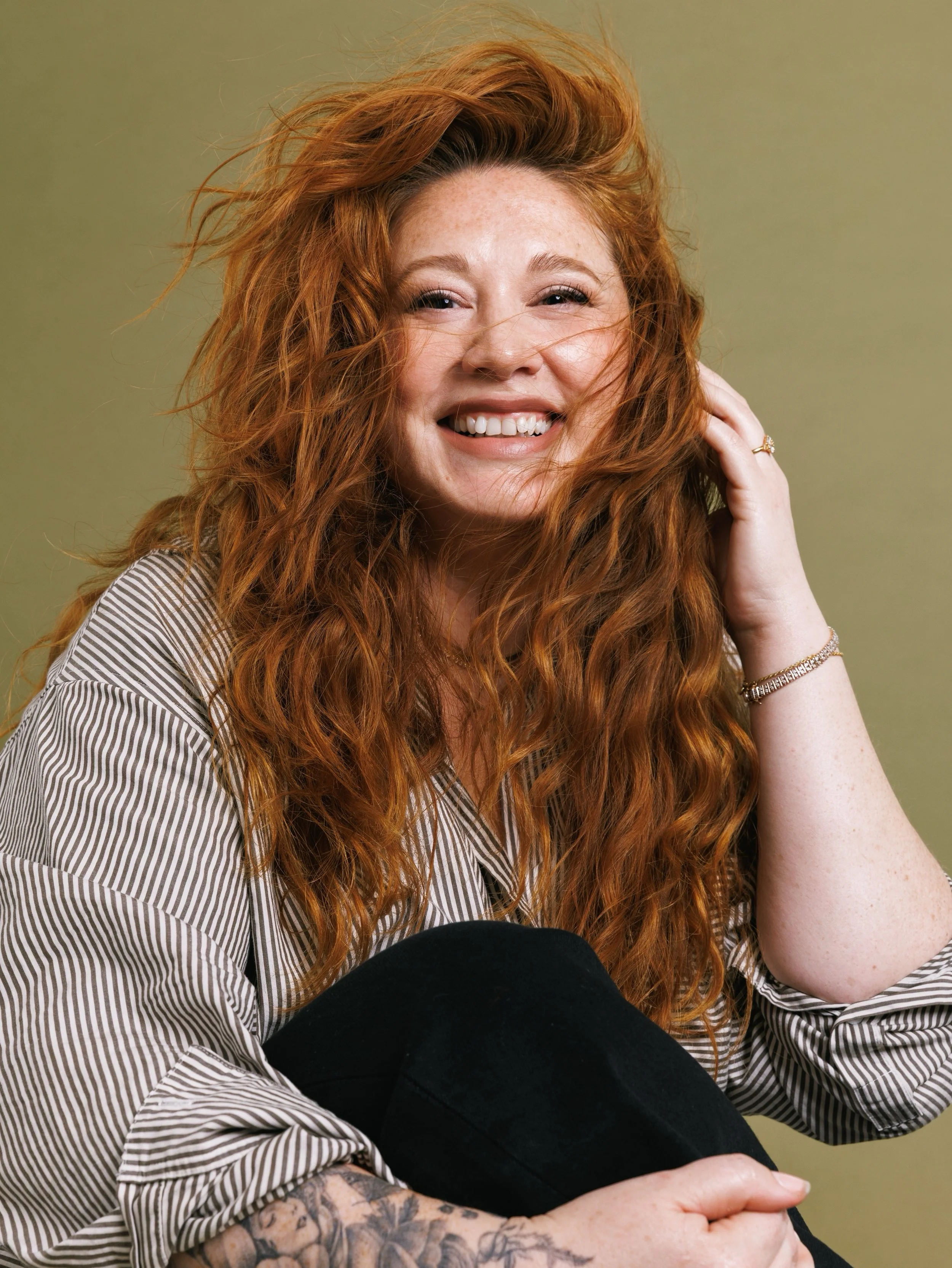 A red-haired woman smiling while sitting with her hand gently touching her face, wearing a striped shirt and jewelry, with someone else's arm and leg visible in the foreground.