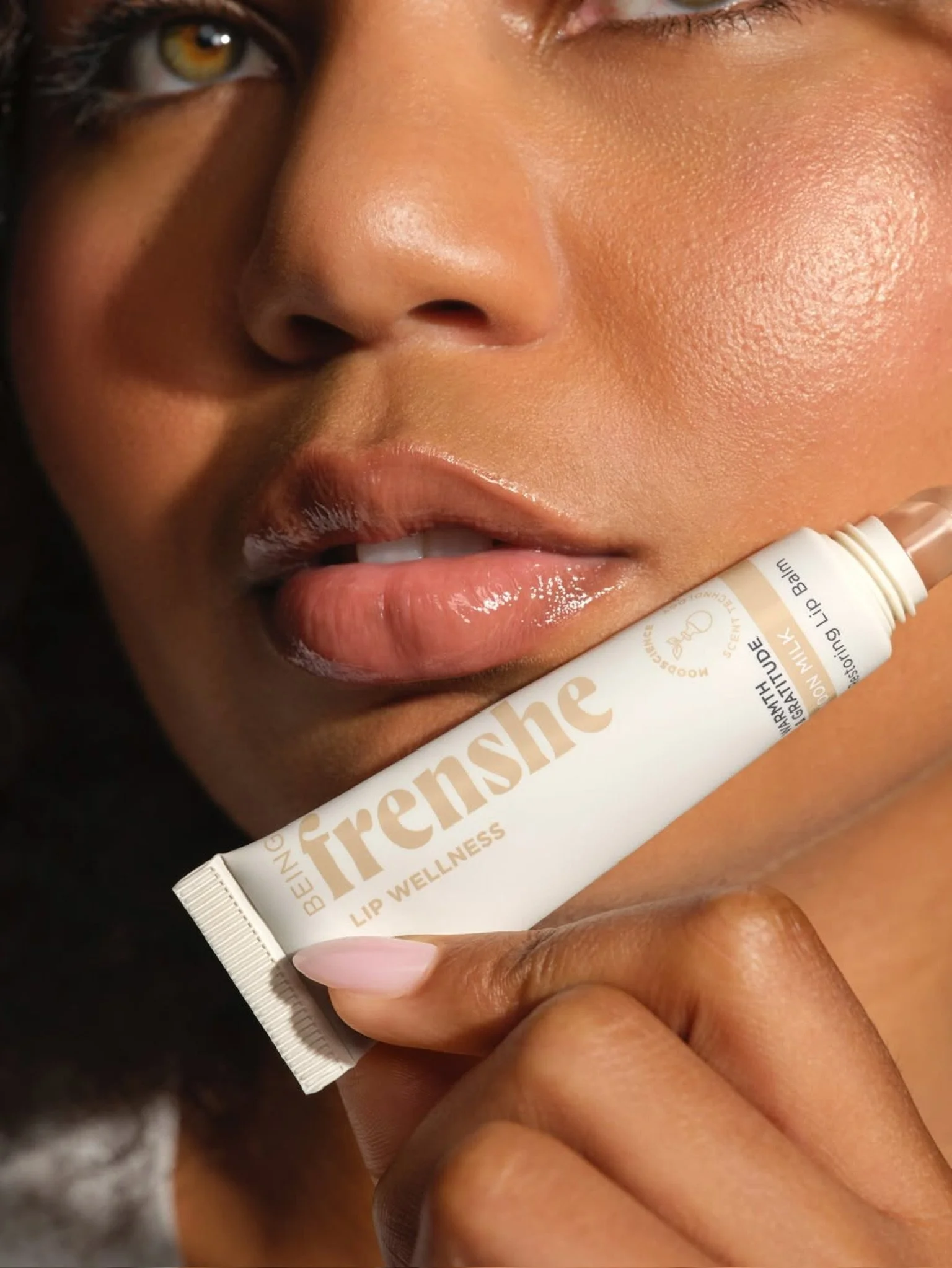 Close-up of a woman holding a lip balm tube near her lips, with her face partially visible, showing her eye, nose, and lips, with smooth skin. Hair and makeup by Amber Rose.