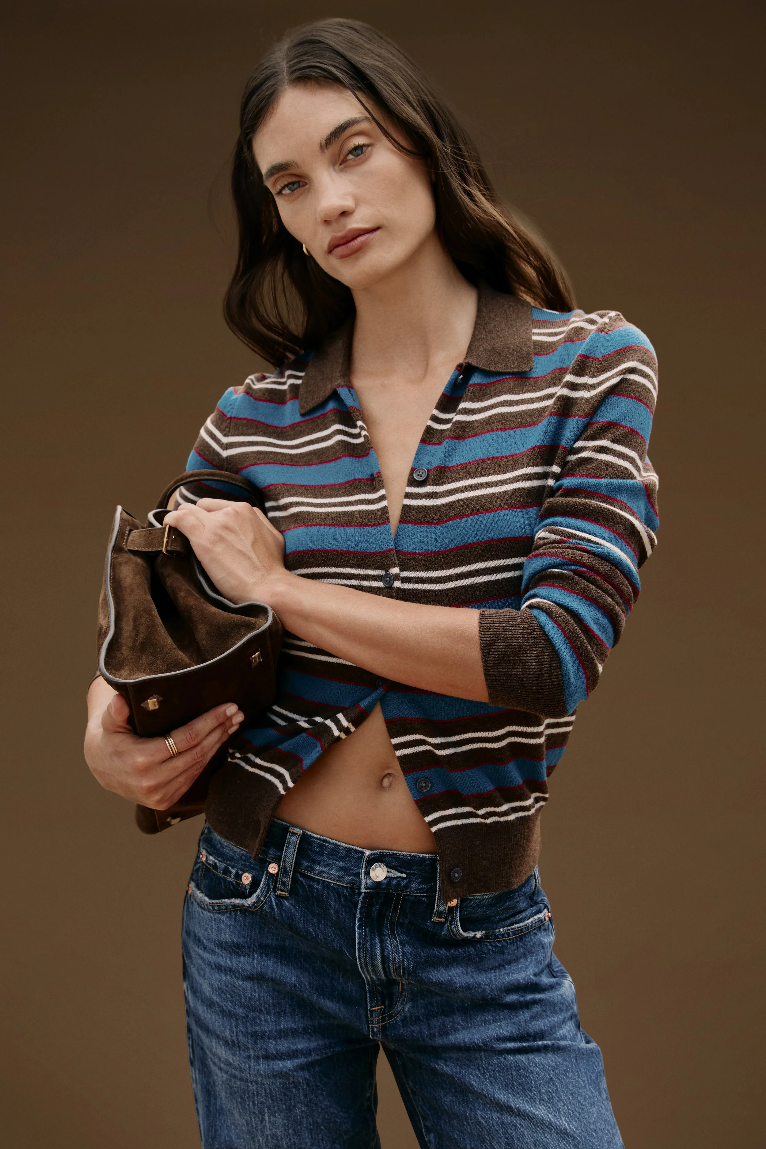 A woman with long dark hair wearing a striped shirt and jeans, holding a brown handbag, with her shirt partially unbuttoned and revealing her midriff. Hair and Makeup by Amber Rose. 
