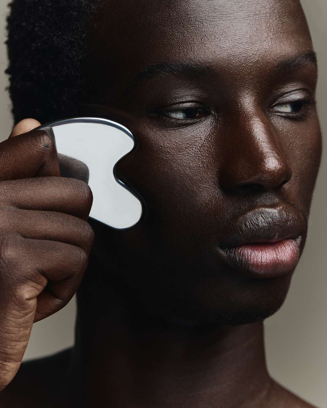 A model with dark skin and short hair using a facial roller on their cheek. Makeup by Amber Rose.
