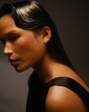 A woman with long dark hair and minimal makeup looking to the side, wearing a black strapless dress against a dark background. Hair and makeup by Amber Rose.