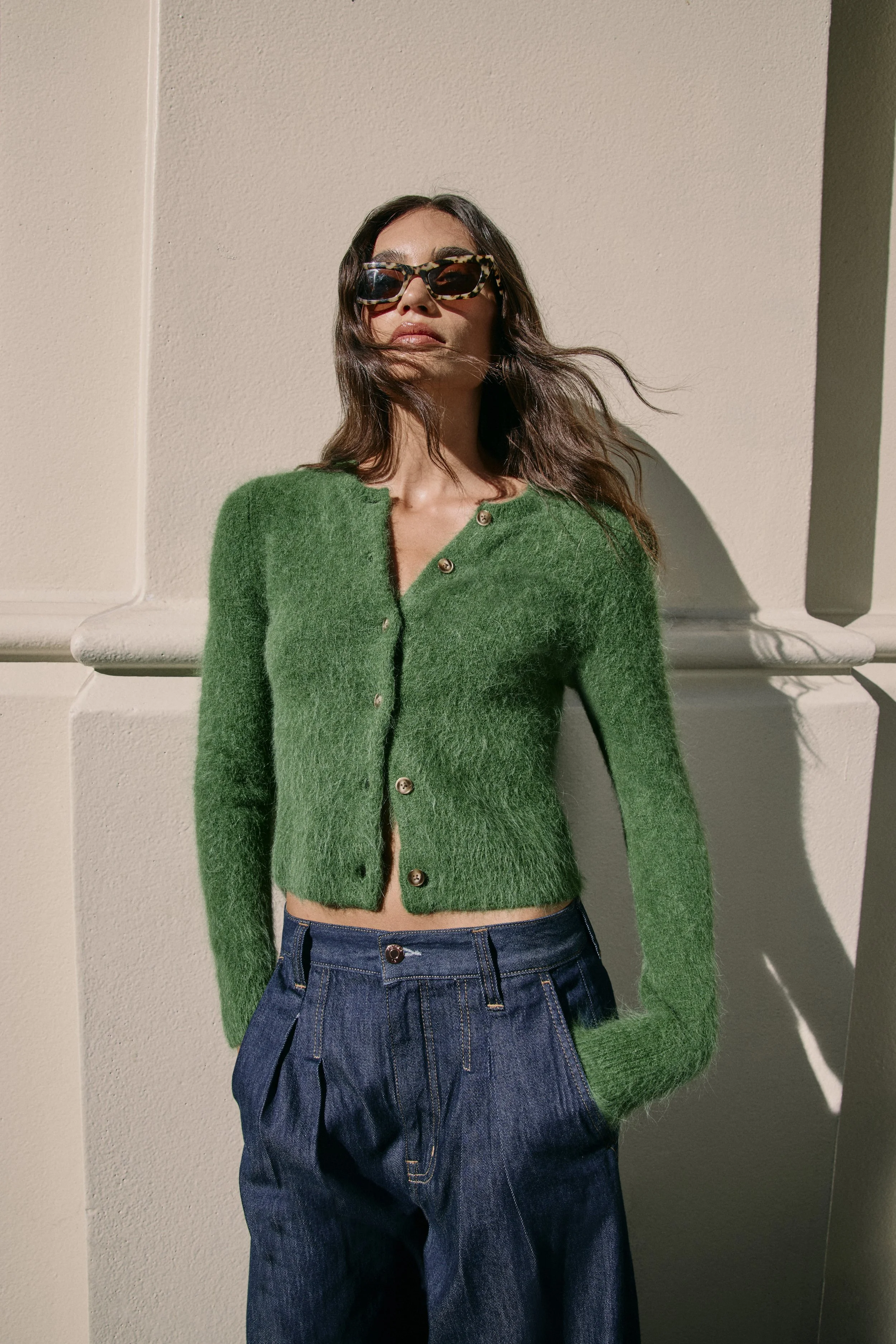 Woman wearing sunglasses, a green cardigan, and dark jeans, standing against a beige wall with shadows.