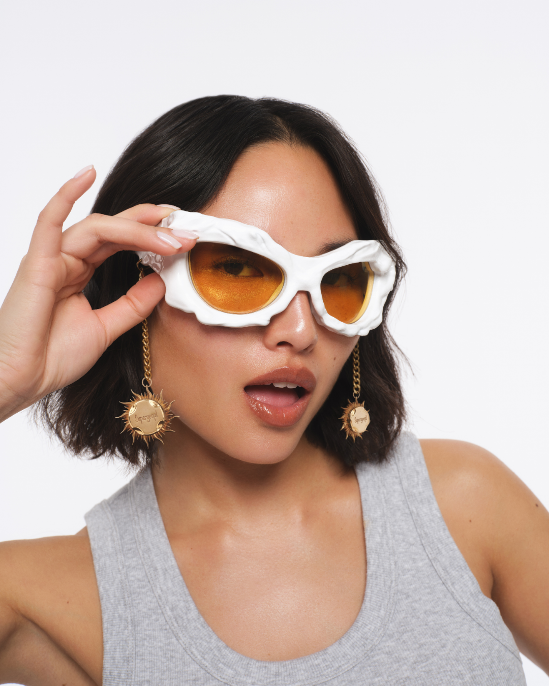 Young woman with dark hair wearing orange-tinted sunglasses with a white shell-shaped frame and large gold earrings, posing with her mouth slightly open against a plain white background. Hair and makeup by Amber Rose.
