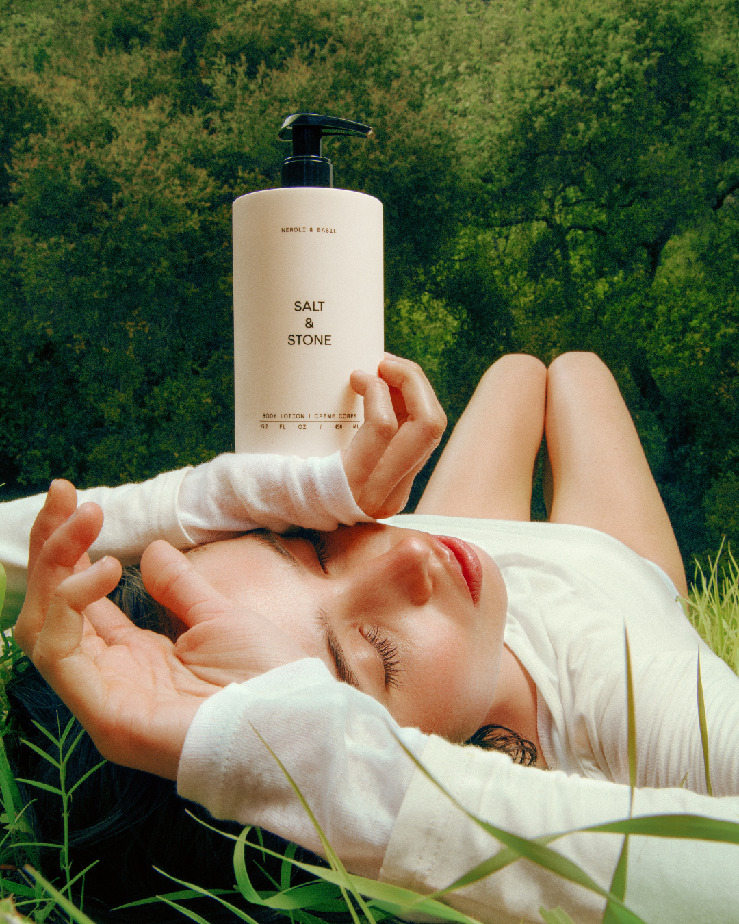A woman lying on the grass in a forest, holding a bottle of body lotion marked 'Salt & Stone'. She appears relaxed with eyes closed, surrounded by greenery.