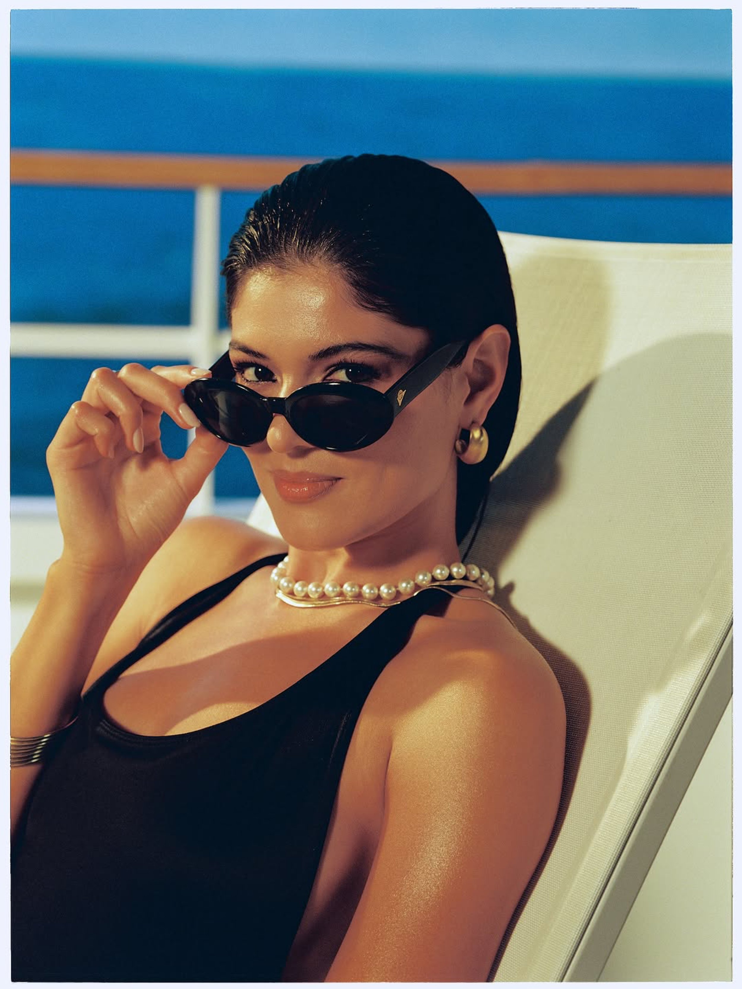 A woman with dark hair and gold earrings is sitting on a lounge chair, peeling back black sunglasses to reveal her eyes, with a blue background and a white chair in the scene. Hair and makeup by Amber Rose.
