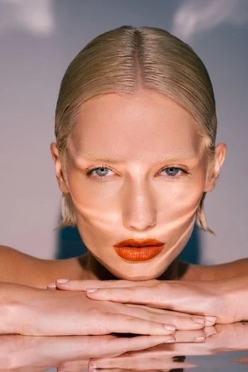 Close-up of a woman with blonde hair, blue eyes, and red lipstick, resting her chin on her folded hands while looking directly at the camera. Los Angeles premiere hair and makeup artist, Amber Rose.