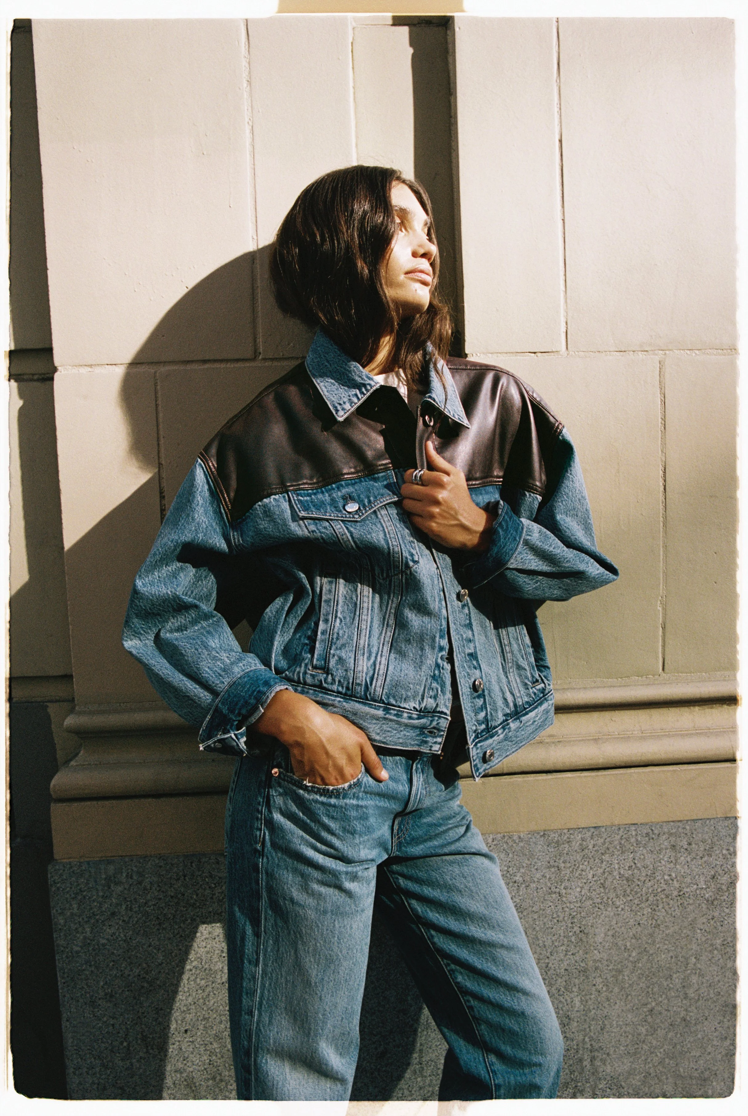 A woman standing against a beige stone wall, wearing a denim and leather jacket and jeans, with her head turned to the side in sunlight.