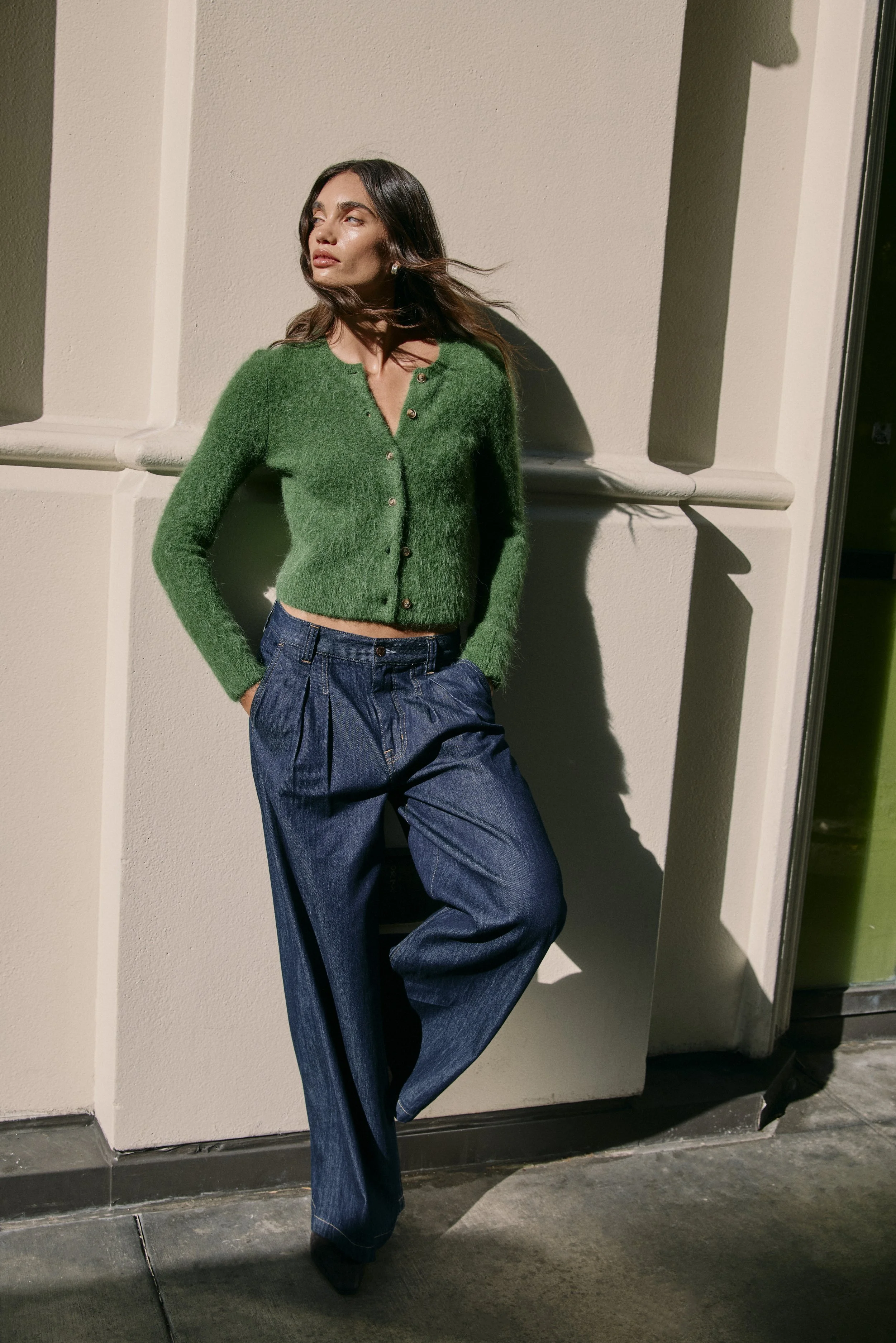 A woman with long brown hair wearing a green cardigan and loose blue jeans, standing against a beige wall with a shadow cast to her side.