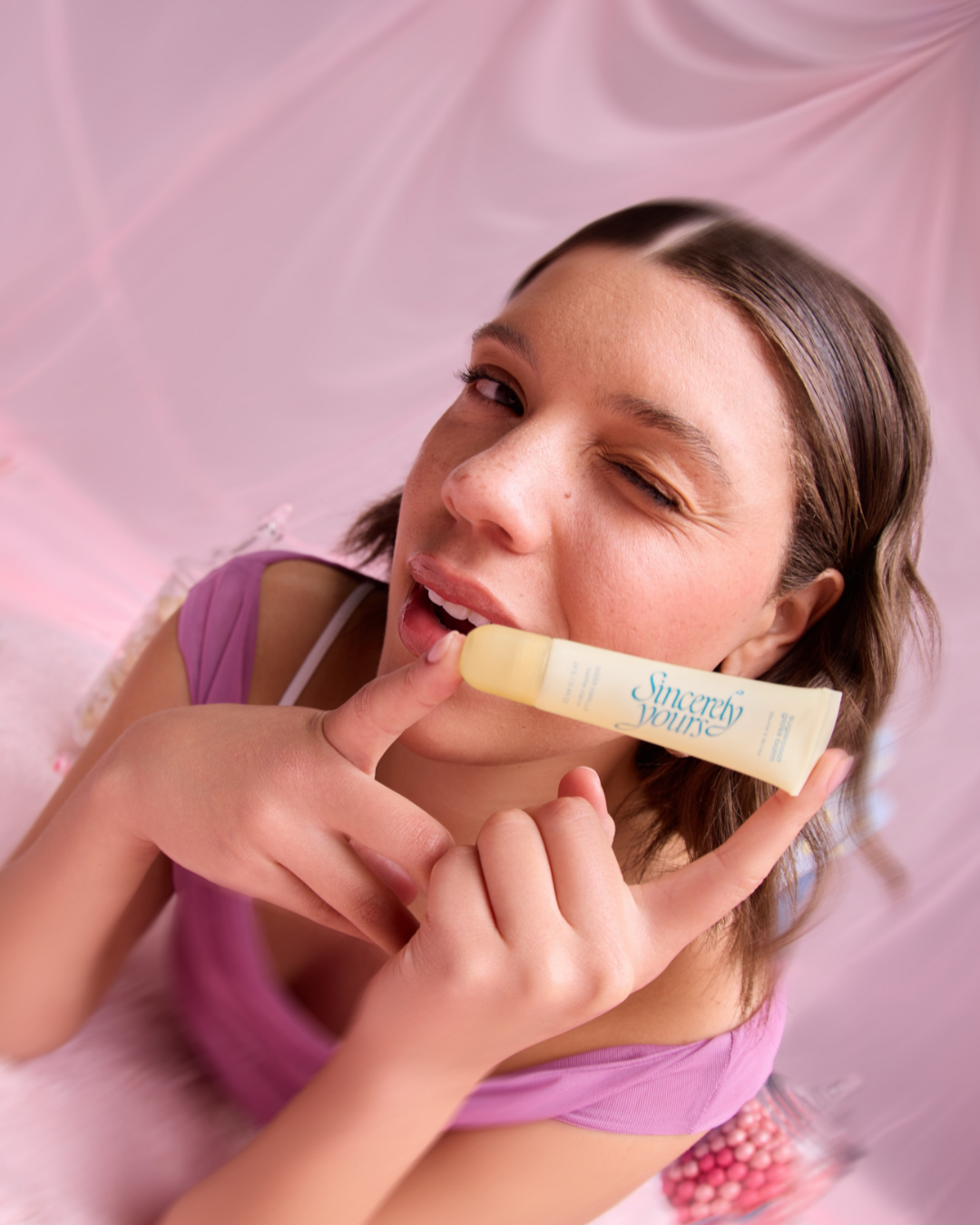 Young woman with short brown hair holding and slightly biting a tube labeled "Sincerely Yours" against a pink background.