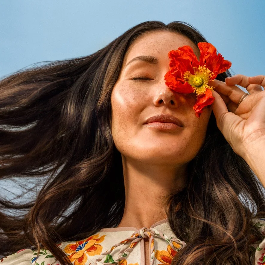 A woman with long, dark hair holding a bright red flower over her eye, against a light blue background.