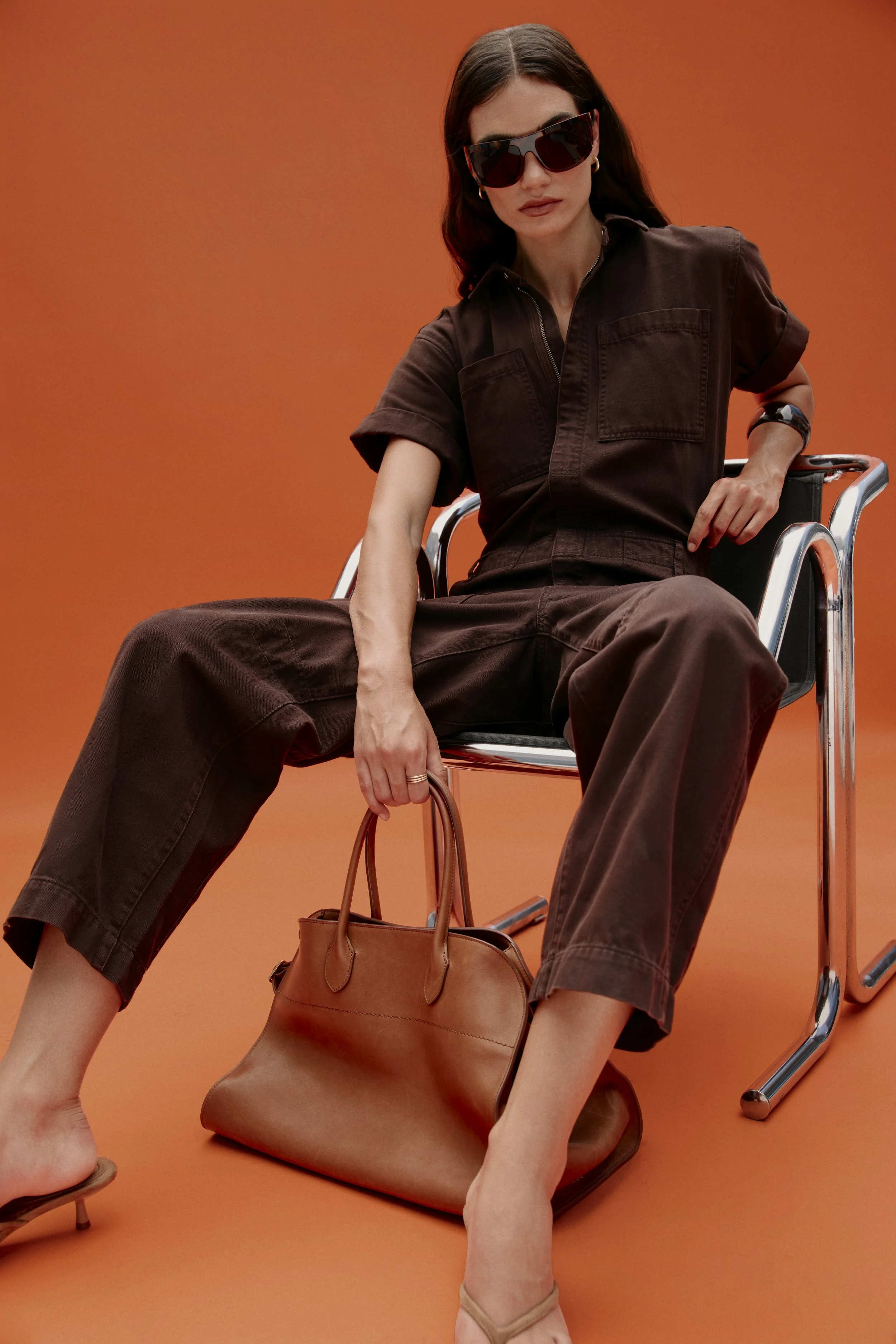A woman sitting in a chair with an orange background, wearing sunglasses, a brown shirt with rolled-up sleeves, and loose brown pants. She is holding a tan handbag and barefoot, with a purse on the floor next to her.