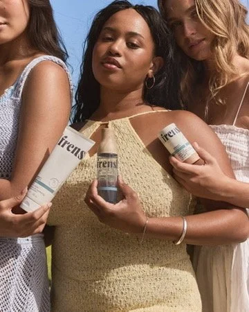 Three women holding bottles of Friends sunscreen outdoors. Hair and makeup by Amber Rose.