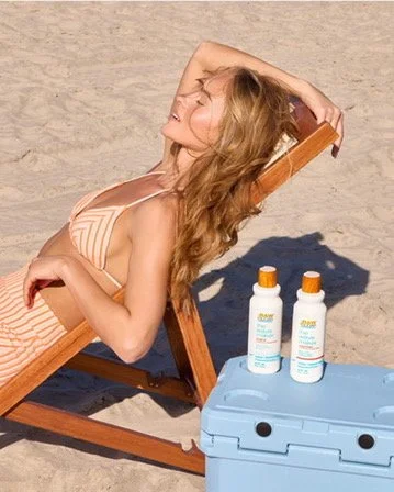 Woman relaxing on a beach lounge chair in a striped bikini, leaning back with her hand behind her head, next to two bottles of sunscreen on a blue cooler.