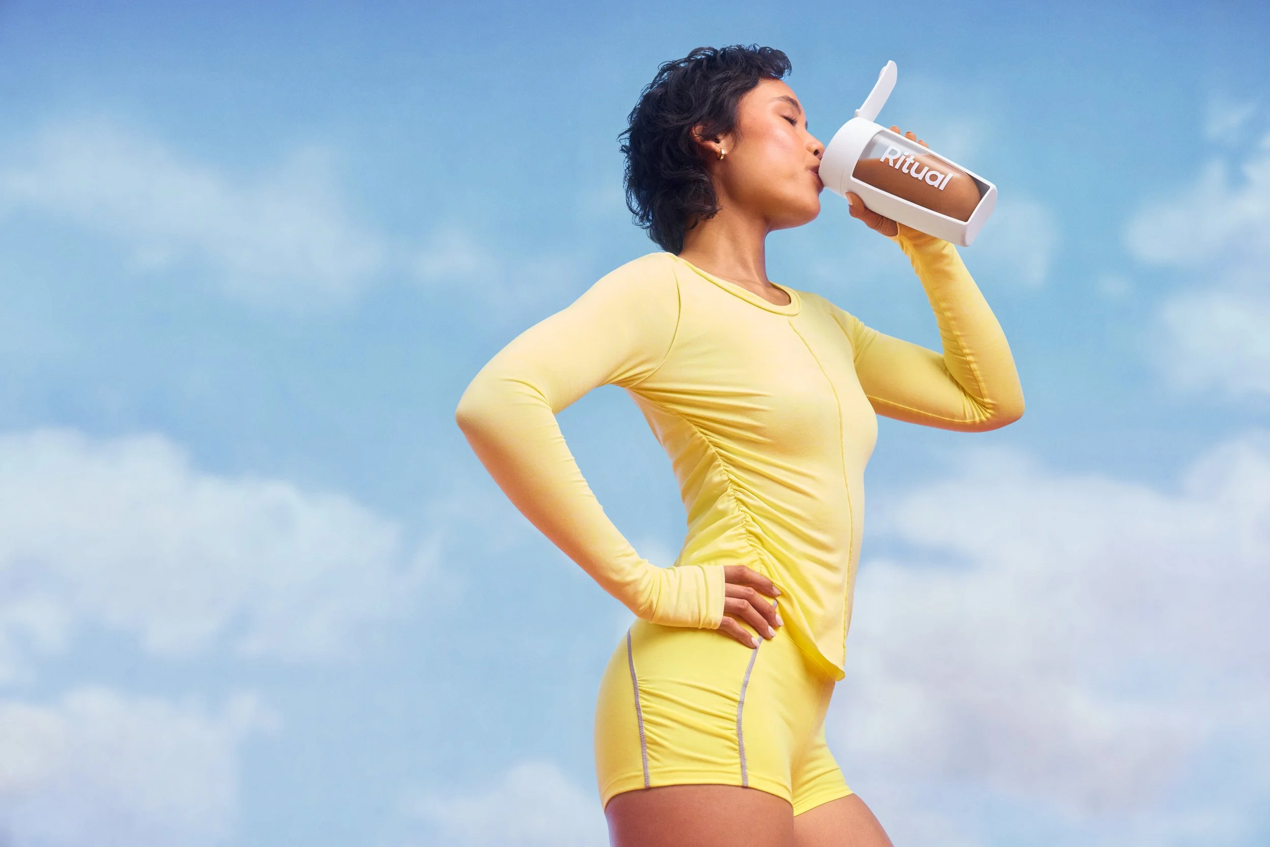 Woman in yellow athletic wear holding a drink labeled 'Ritual' and drinking it, standing outdoors against a blue sky with a few clouds.