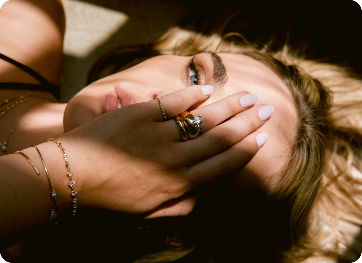 A woman with blonde hair and blue eyes lying down, covering part of her face with her hand that has rings and bracelets. Hair and makeup by Amber Rose.