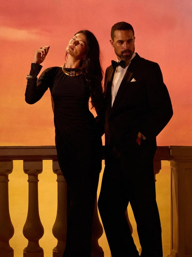 A woman and a man dressed in formal evening attire standing on a balcony with a sunset sky in the background. Hair and makeup by Amber Rose.