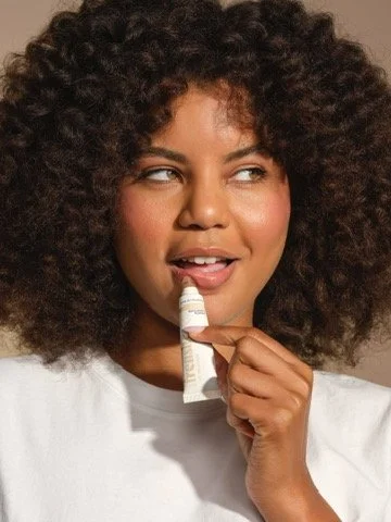A woman with large curly hair applying lip balm. Hair and makeup by Amber Rose.