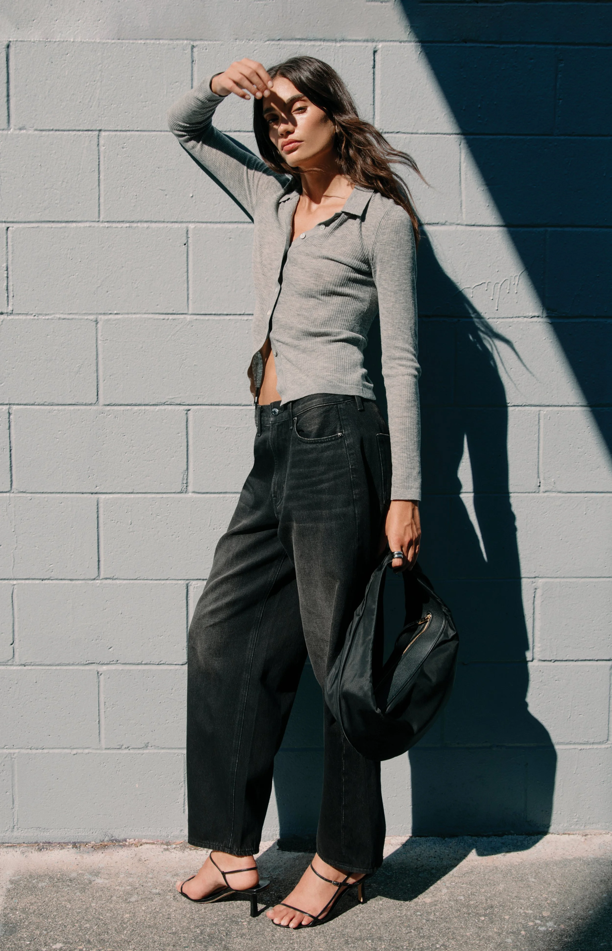 A woman with long dark hair wearing a gray button-down top and black wide-leg jeans standing against a light gray cinder block wall, shielding her eyes from the sun with one hand, holding a black bag, and wearing black strappy high heels.