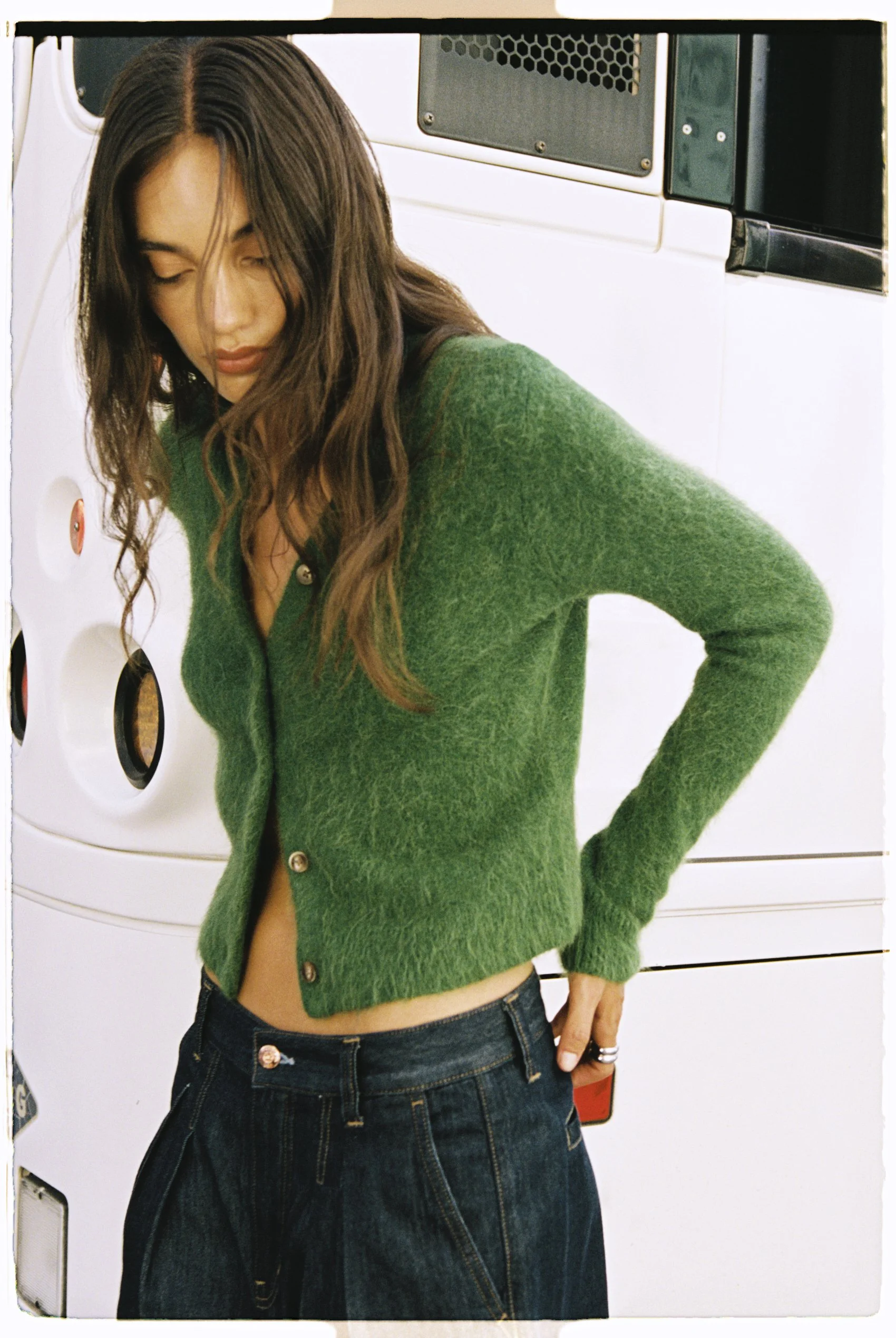 A woman with long wavy brown hair wearing a green cardigan shirt and denim jeans standing in front of a white laundry machine with black and red accents.