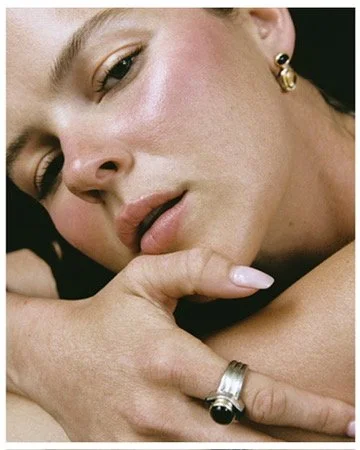 Close-up beauty portrait with chin resting on a hand stacked with statement rings, dewy blush skin, jewelry editorial by Los Angeles makeup artist Amber Rose.