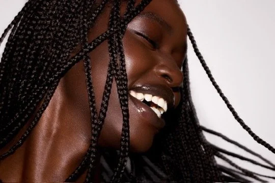 Close-up of a smiling Black woman with braided hair, showing her teeth and dimples. Los Angeles premiere hair and makeup artist, Amber Rose.