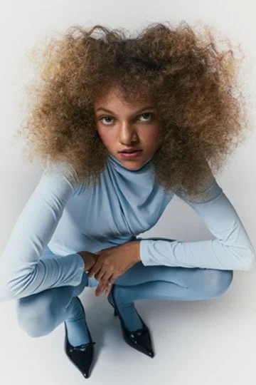 Woman with voluminous curly hair wearing a light blue outfit and black heels, squatting against a white background. Hair and makeup by Amber Rose.