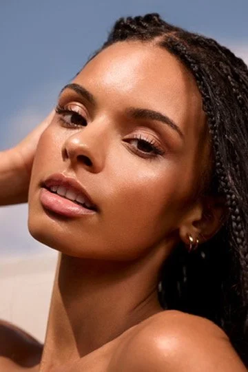 Close-up of a young woman with long braided hair, natural makeup, and earrings, posing outdoors against a blue sky. Hair and makeup by Amber Rose.