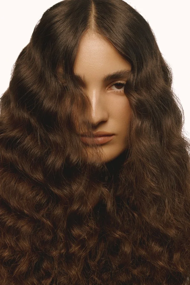A young woman with long, thick, curly brown hair covering half of her face, wearing a neutral expression against a plain white background. Hair and makeup by Amber Rose.