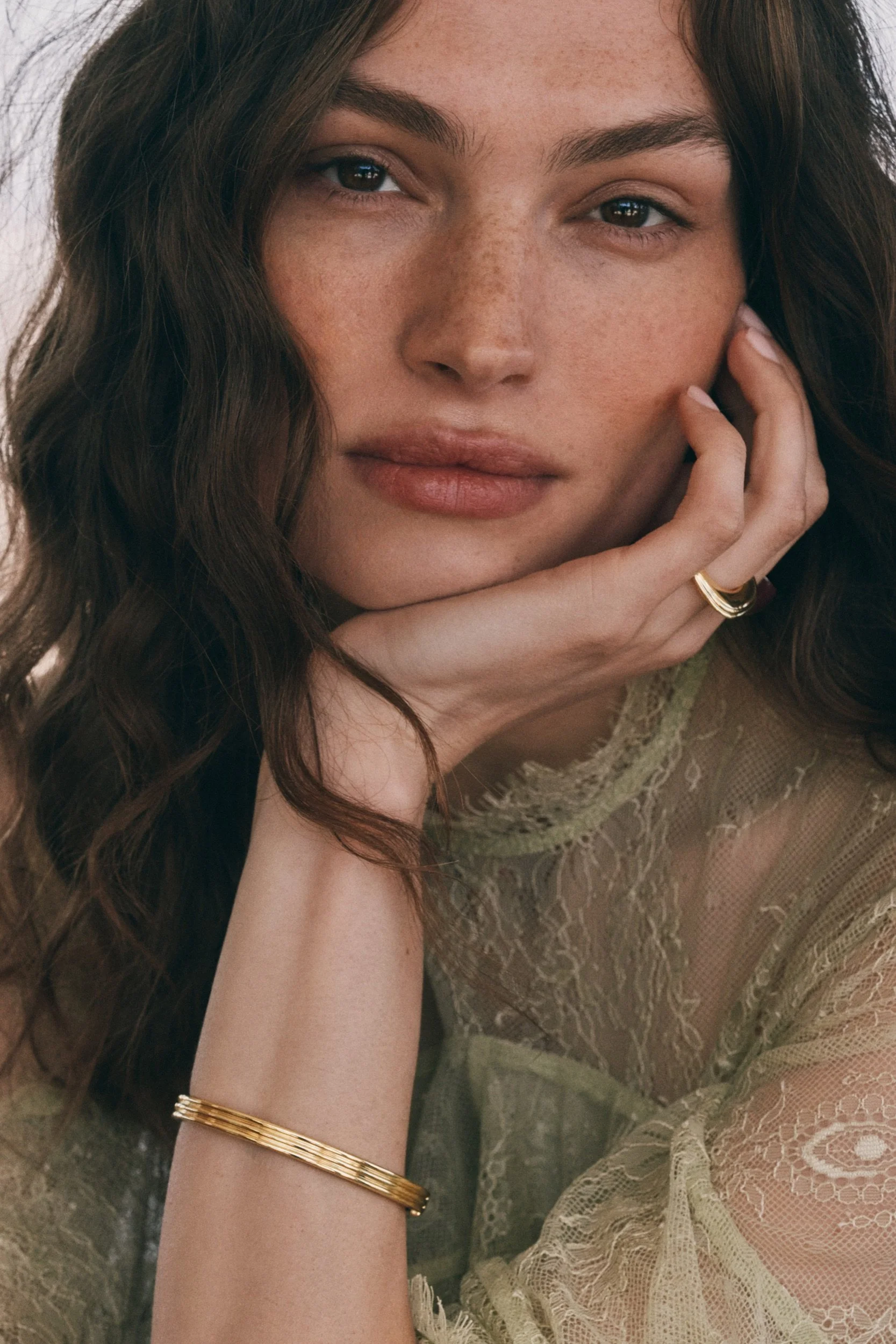 A woman with long, wavy brown hair and freckles, resting her head on her hand, wearing a sheer, lace, beige dress, gold rings, and a gold bracelet.