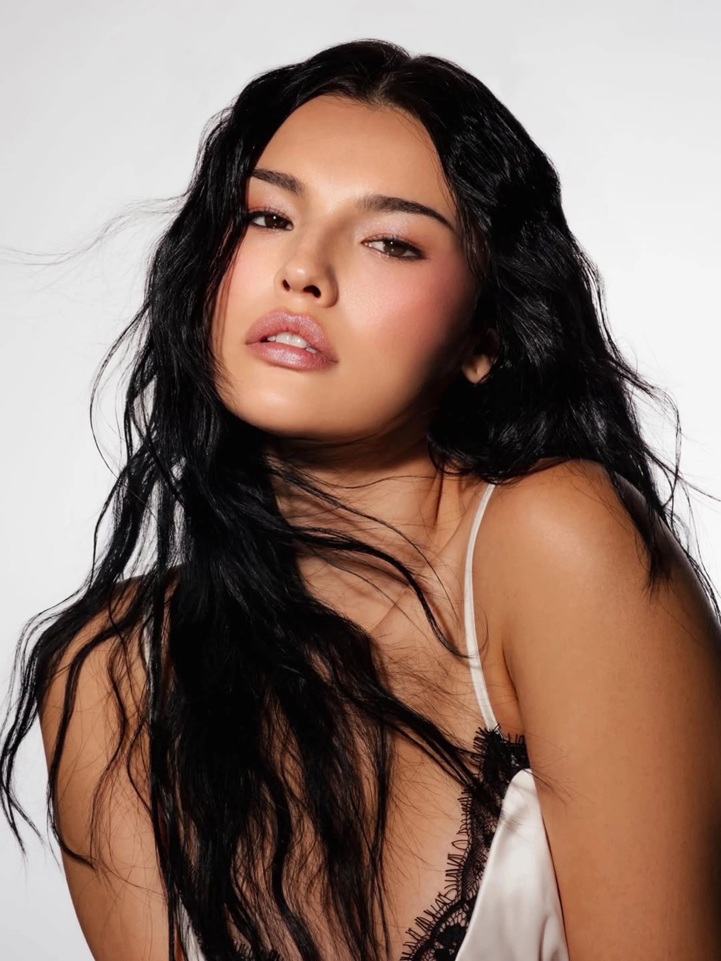 A woman with long black hair, wearing a white satin top with black lace detailing, looking with a relaxed expression against a plain light background. Hair and makeup by Amber Rose.