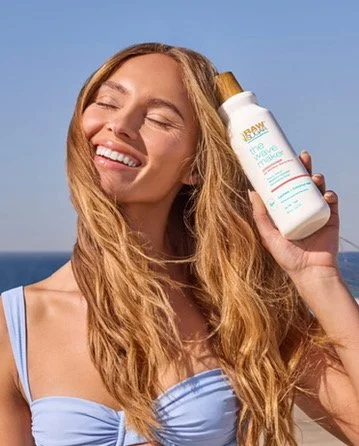 Smiling woman with long wavy hair holding a bottle of lotion outdoors