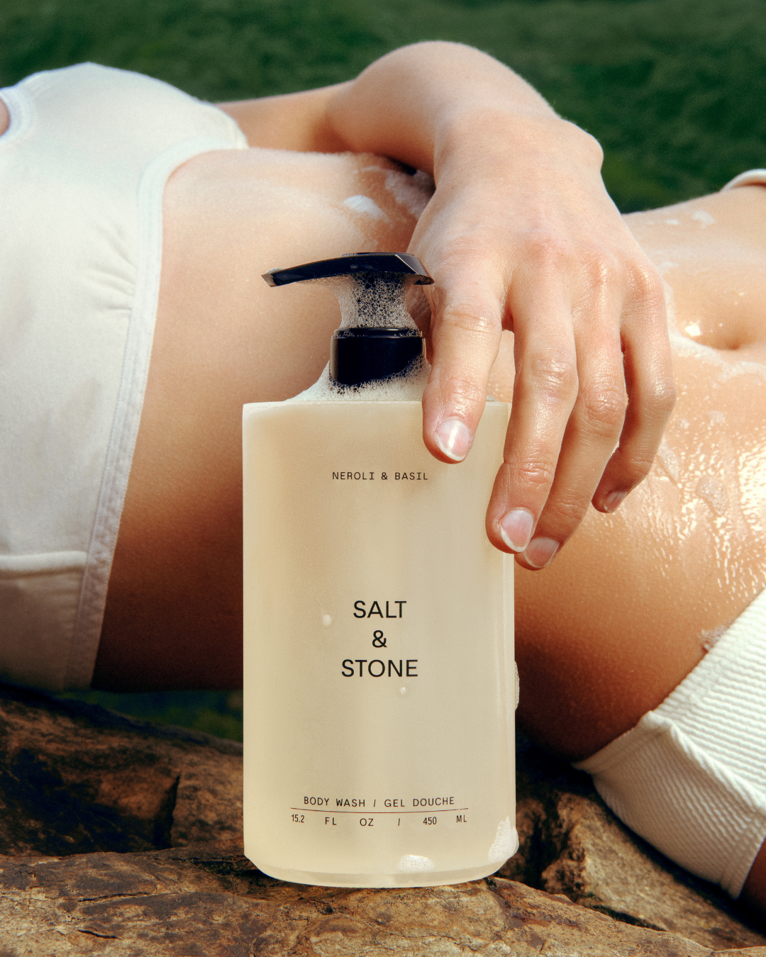 A person lying on a rock outdoors, holding a bottle of Neroli & Basil Salt & Stone body wash with foam, wearing a white outfit and with one hand resting on the ground.