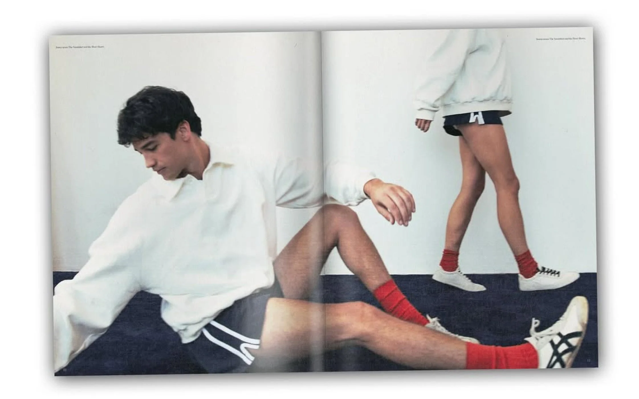 Two young men with athletic builds wearing white sweatshirts, black shorts, red socks, and sneakers, engaging in a dance move or pose in a plain indoor setting. Hair and makeup by Amber Rose.