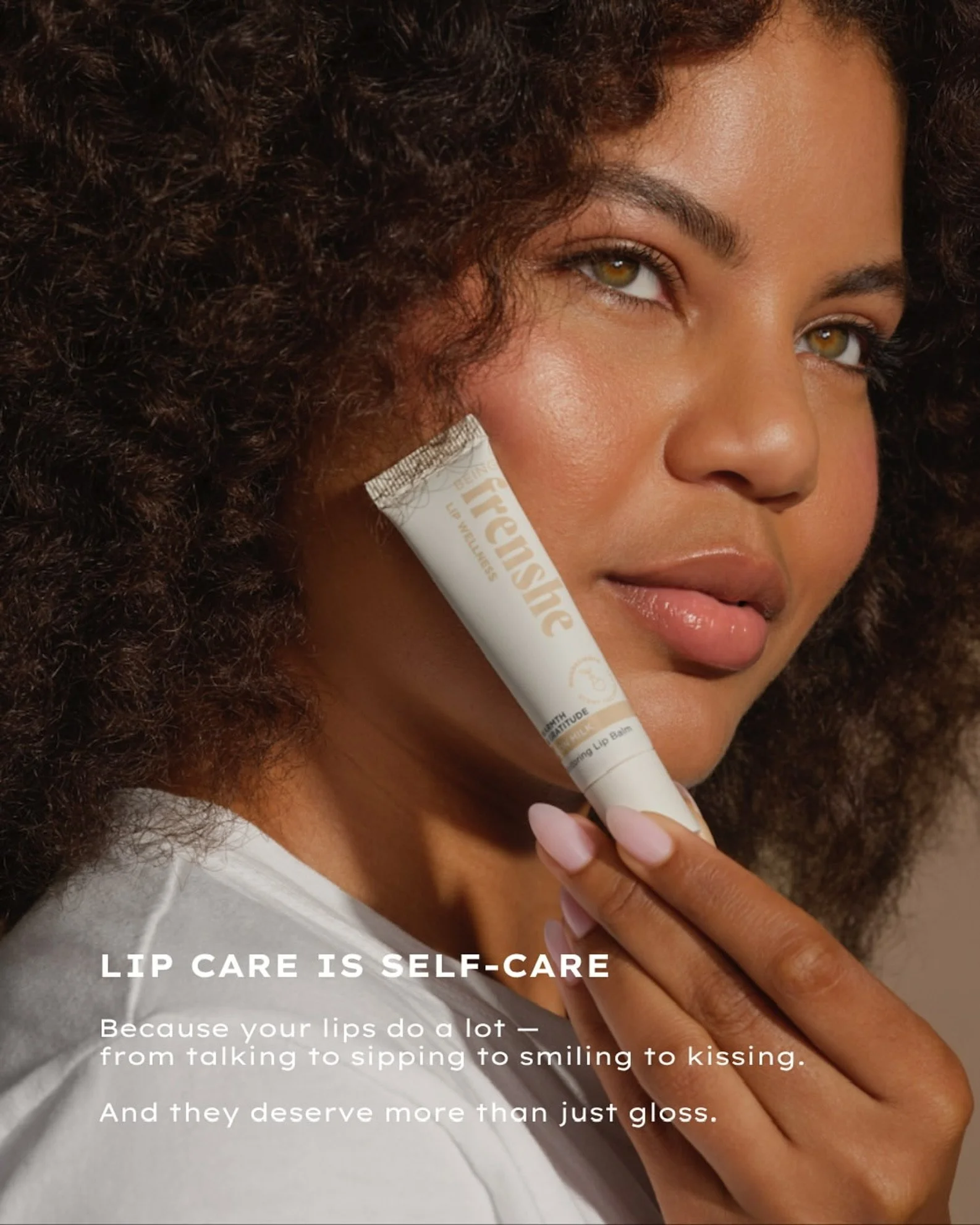 Close-up of a woman with curly hair holding a tube of lip balm to her lips, with the text overlay reading 'Lip Care is Self-Care. Because your lips do a lot — from talking to sipping to smiling to kissing. And they deserve more than just gloss.'. Hai
