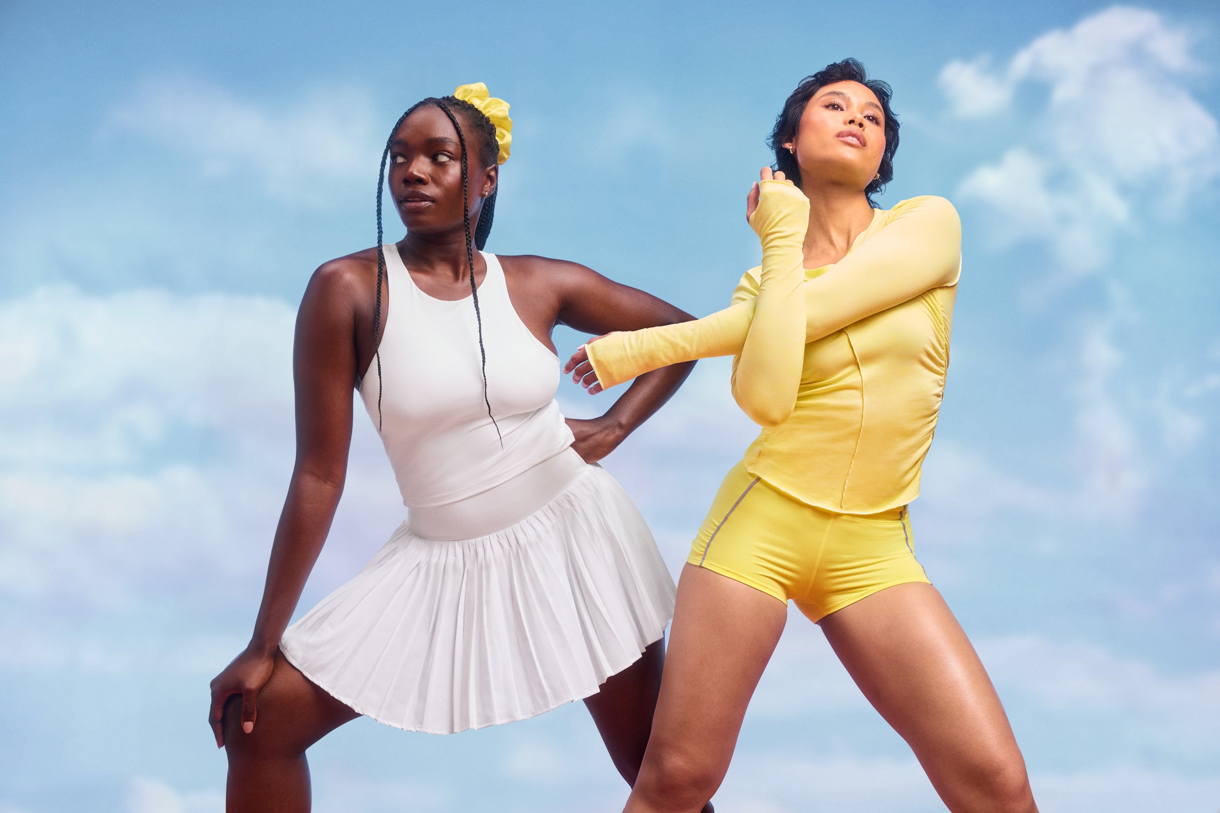 Two women standing outdoors against a blue sky with clouds. One woman is wearing a white top with a white pleated skirt and a yellow scrunchie in her braided hair. The other woman is wearing a yellow long-sleeve top and yellow shorts, with short blac