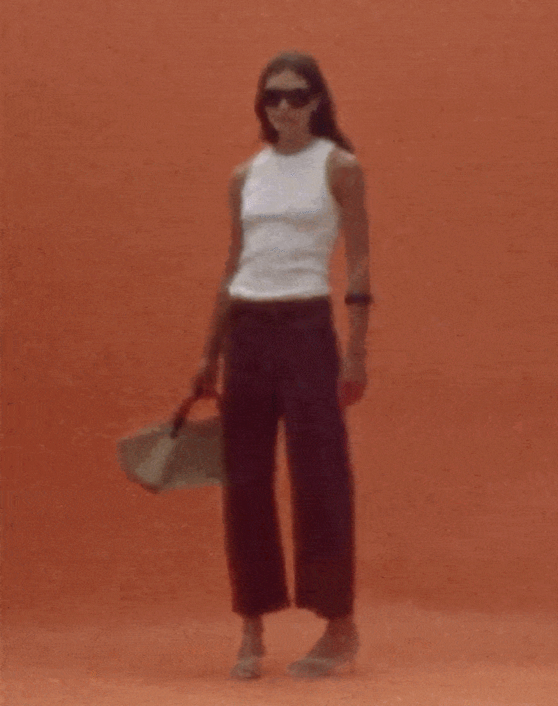 A woman in sunglasses, sleeveless white top, dark wide-leg pants, and high heels, standing against an orange background and carrying a handbag.