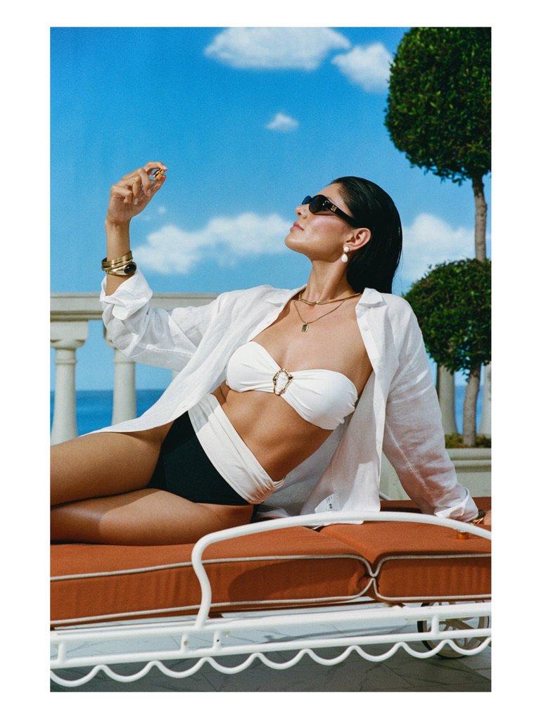 A woman relaxing on a beach chair outdoors, wearing sunglasses, a white strapless bikini top, black swimsuit bottoms, a white shirt, and jewelry, with a bright blue sky, some clouds, and green trees in the background. Hair and makeup by Amber Rose.