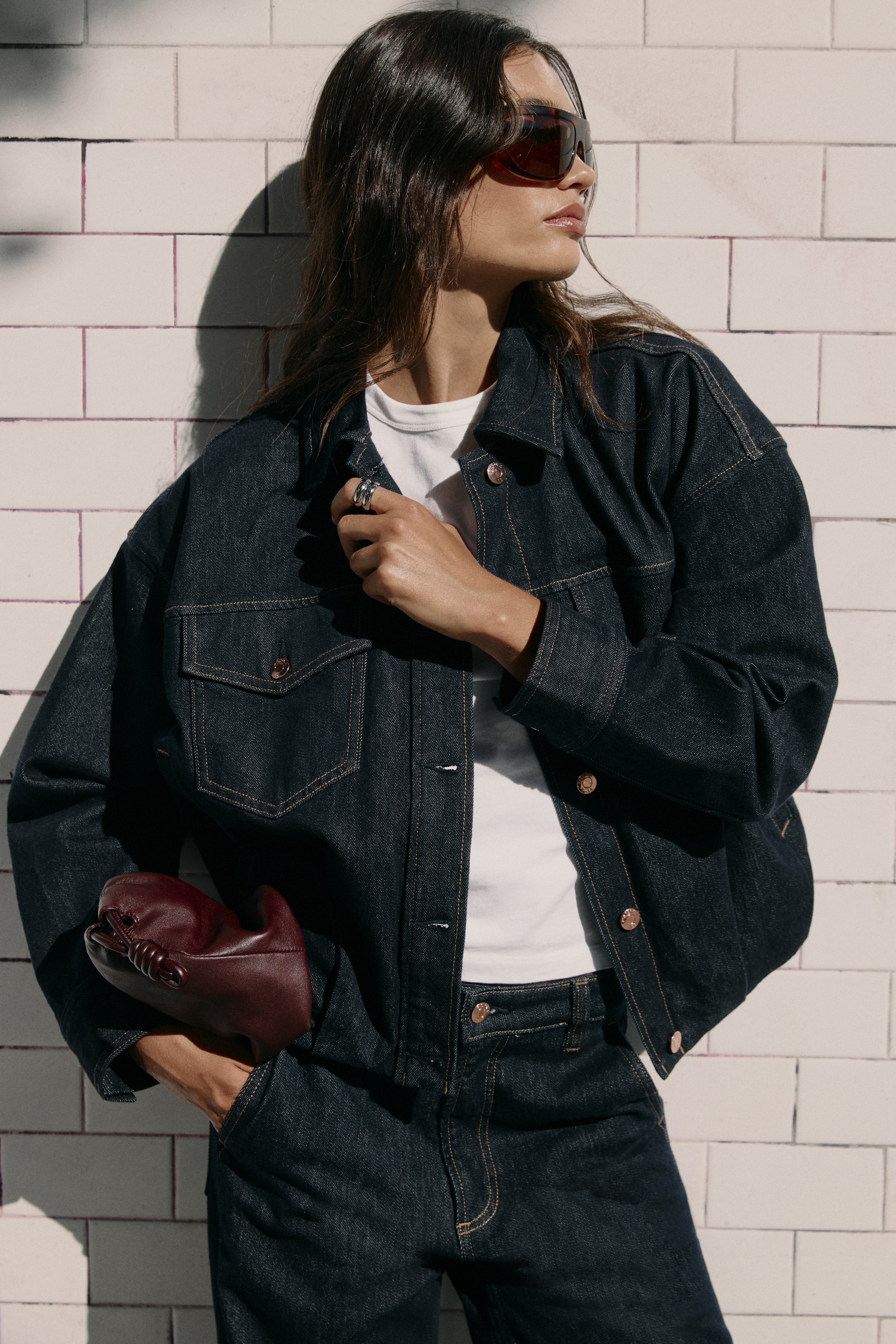 A woman with long dark hair wearing sunglasses, a white t-shirt, and a denim jacket stands against a white brick wall. She holds a red bag and has her hand on her chest, looking to the side.