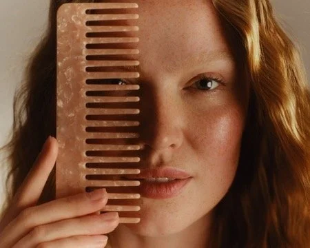 Portrait of a young woman with red hair holding a pink hair comb against half of her face. Hair and makeup by Amber Rose.
