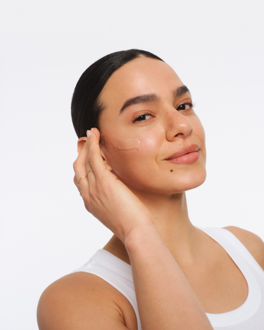 A woman with dark hair and clear skin, wearing a white sleeveless top, applying skincare product to her face against a white background. Hair and makeup by Amber Rose.