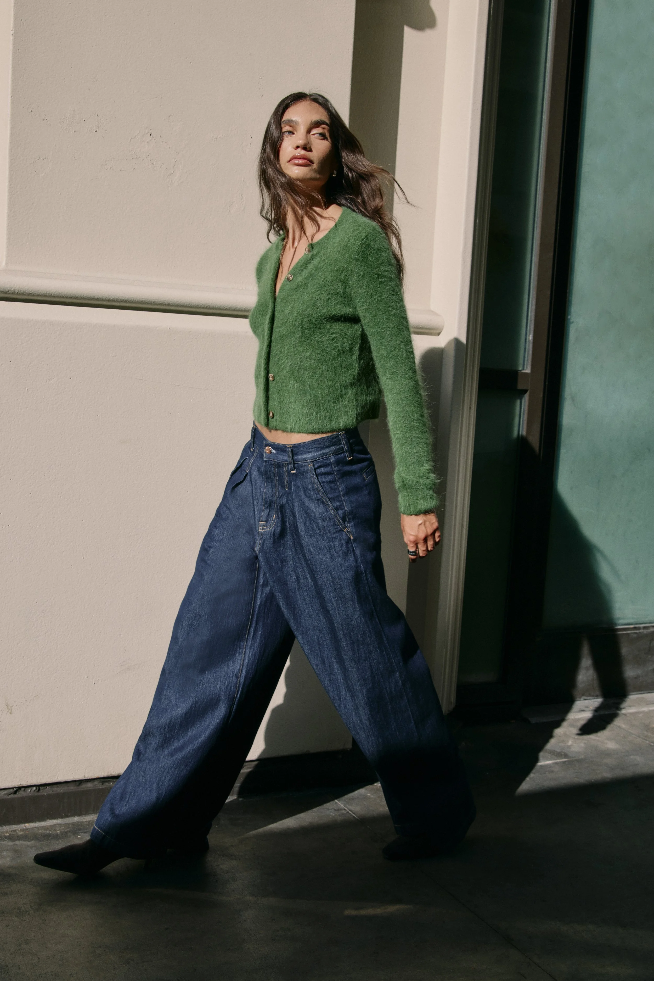 A woman standing outdoors near a building, wearing a green cardigan, wide-leg jeans, and black boots, with a shadow cast on the wall behind her.